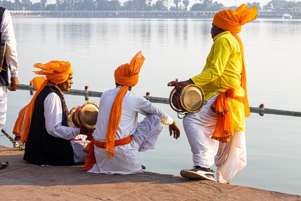 Three men in traditional attire play music by a serene riverside, showcasing Indian culture.